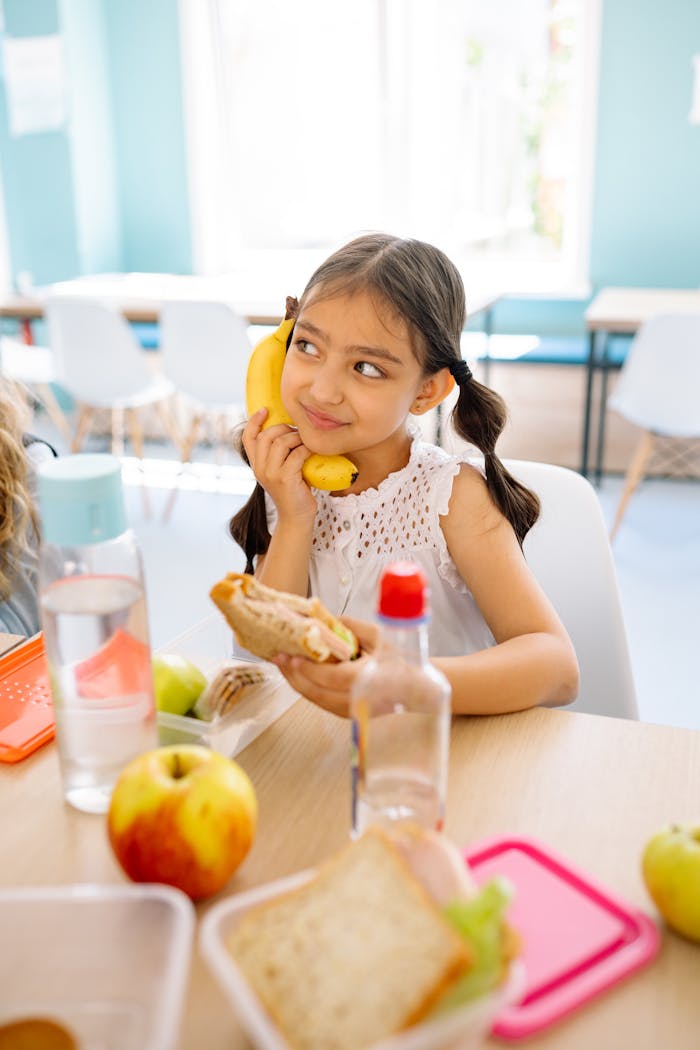 A young girl playfully holds a banana while enjoying a sandwich at a classroom table.